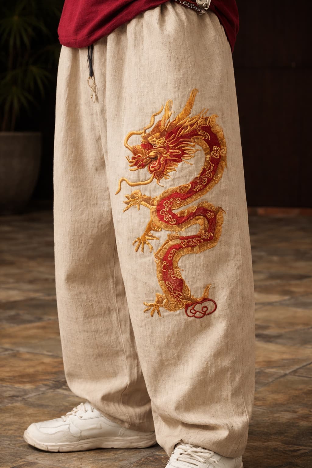Unisex model wearing loose-fitting beige oriental streetwear pants with a large red dragon motif styled with classic white sneakers on a tiled floor.