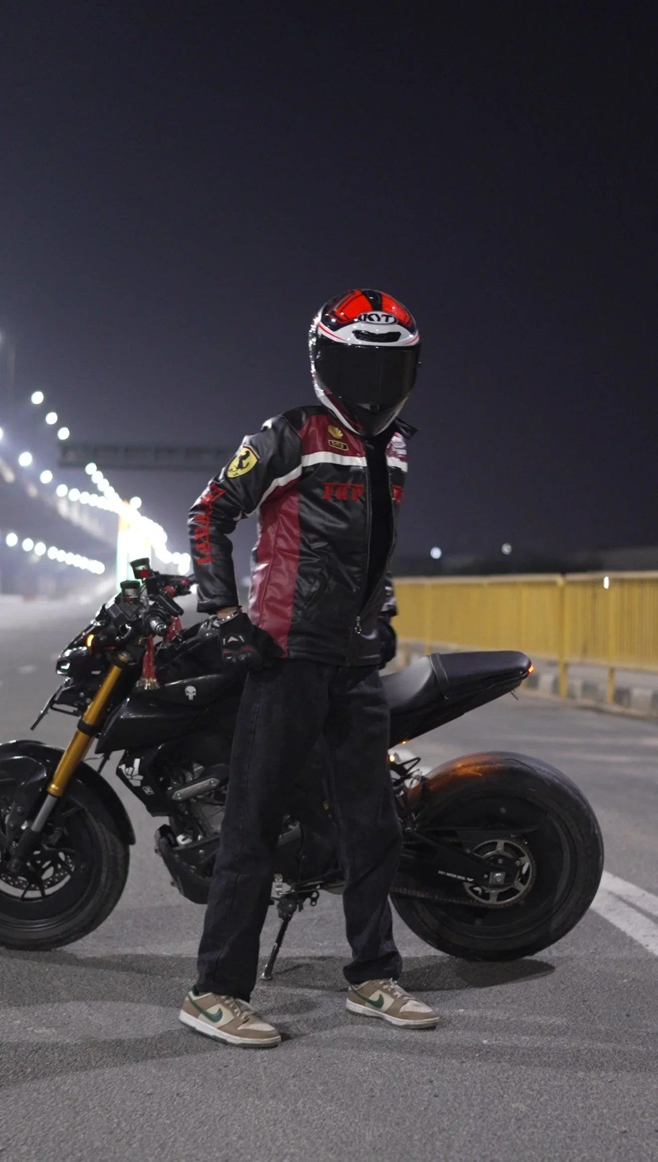 Motorcyclist in reinforced shoulder streetwear jacket and helmet posing by black motorcycle at night
